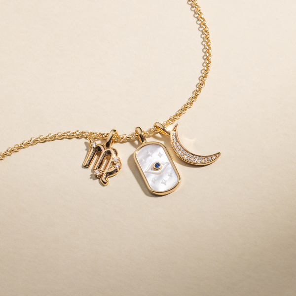 A gold chain necklace featuring a Virgo zodiac symbol charm, an oval charm with an eye design and white inlay, and a crescent moon charm covered in clear gemstones.