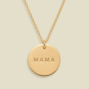 A gold round pendant necklace engraved with the word 'MAMA' in capital letters, suspended from a delicate gold chain against a solid light beige background.