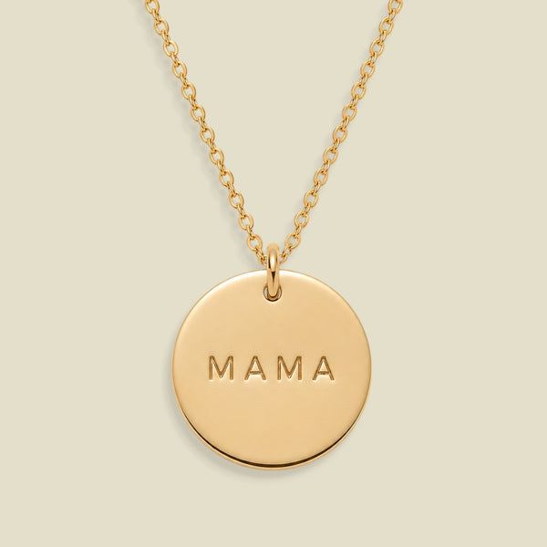 A gold round pendant necklace engraved with the word 'MAMA' in capital letters, suspended from a delicate gold chain against a solid light beige background.