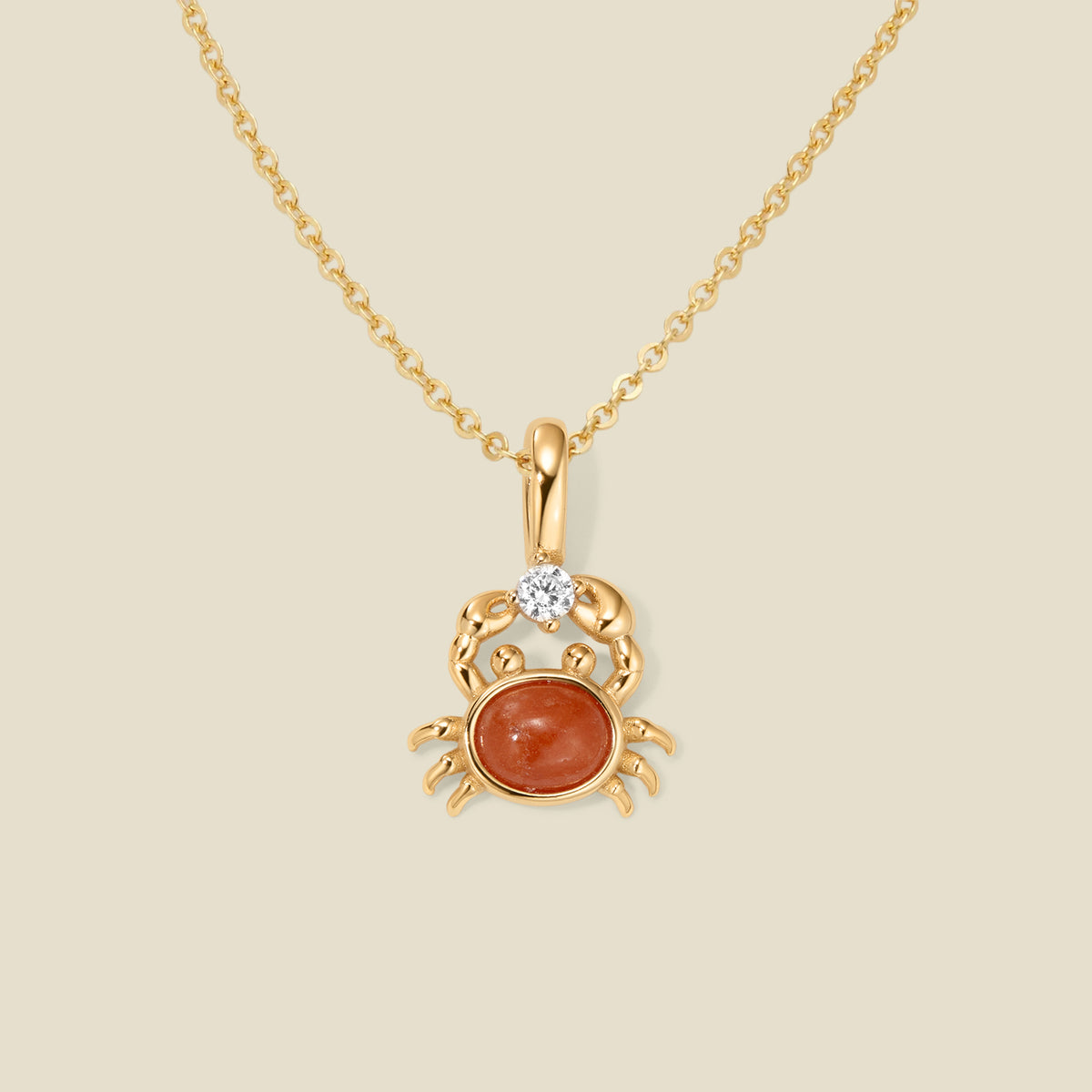 A dainty gold chain necklace with a gold crab-shaped pendant with a red stone and clear accent stone on a beige background