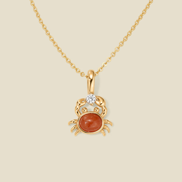 A dainty gold chain necklace with a gold crab-shaped pendant with a red stone and clear accent stone on a beige background