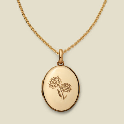 A dainty gold chain necklace with an oval locket pendant, featuring an engraved floral motif on the front, set against a solid beige background.