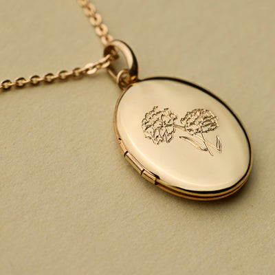 A close up shot of a gold oval locket, engraved with a floral design on the front and laid on a textured olive green background.