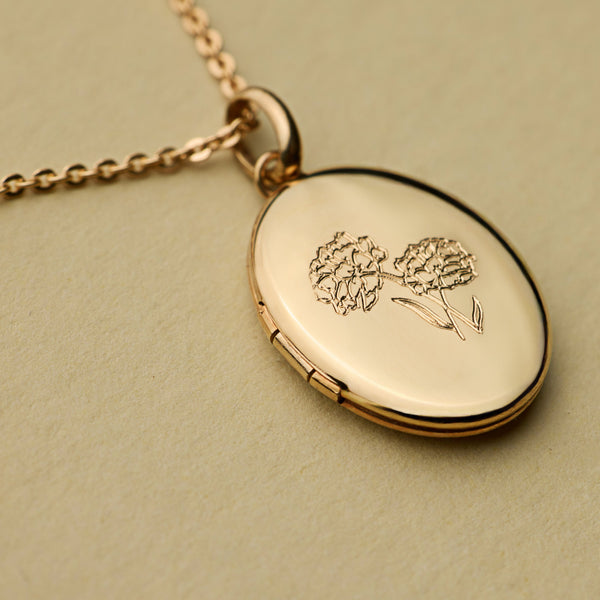 A close up shot of a gold oval locket, engraved with a floral design on the front and laid on a textured olive green background.