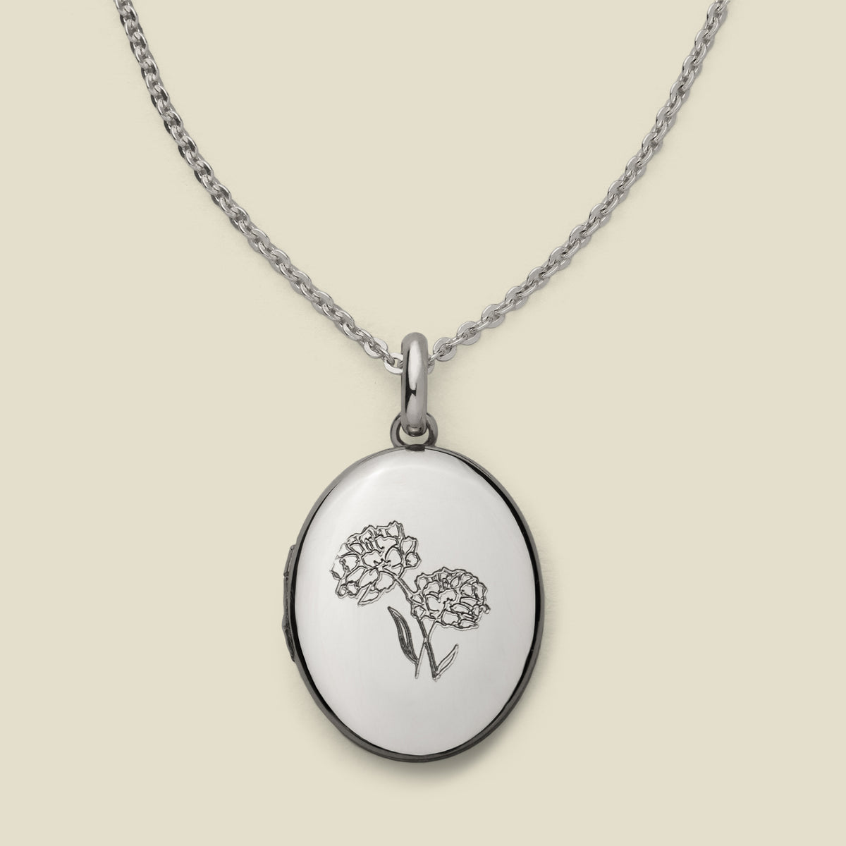 A dainty silver chain necklace with an oval locket pendant, featuring an engraved floral motif on the front, set against a solid beige background.