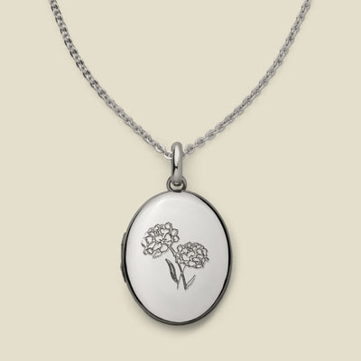 A dainty silver chain necklace with an oval locket pendant, featuring an engraved floral motif on the front, set against a solid beige background.