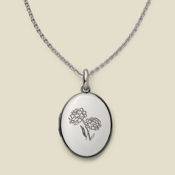 A dainty silver chain necklace with an oval locket pendant, featuring an engraved floral motif on the front, set against a solid beige background.