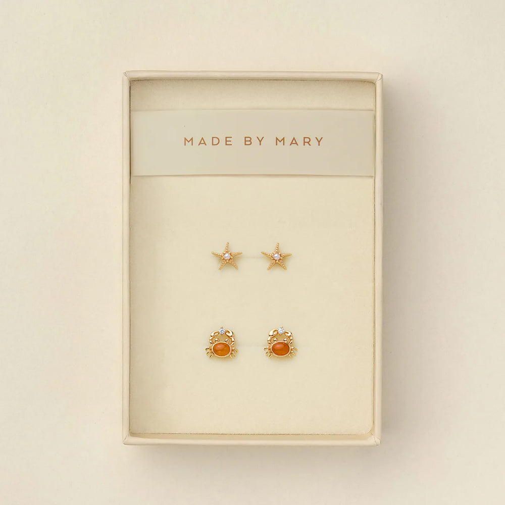 A pair of gold starfish stud earrings with pearls in the center, and a pair of gold crab stud earrings with amber-orange centers and clear crystals, presented in a cream-colored jewelry box with 'MADE BY MARY' printed on the inside header.