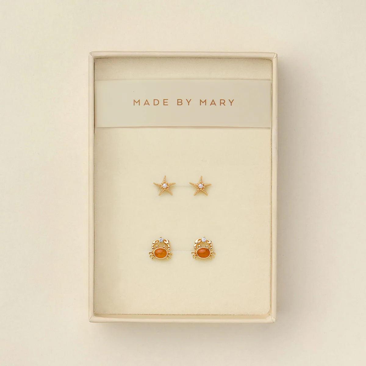 A pair of gold starfish stud earrings with pearls in the center, and a pair of gold crab stud earrings with amber-orange centers and clear crystals, presented in a cream-colored jewelry box with 'MADE BY MARY' printed on the inside header.