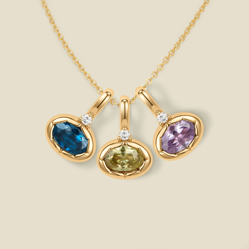 A dainty gold chain necklace with three oval pendant charms, a blue-green, light green, and light purple oval gemstone on each with a small clear accent stone above them, set against a solid beige background.