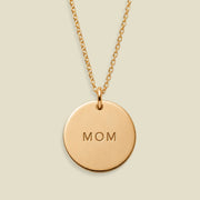 A gold round pendant necklace engraved with the word 'MOM', suspended from a delicate gold chain against a light beige background.