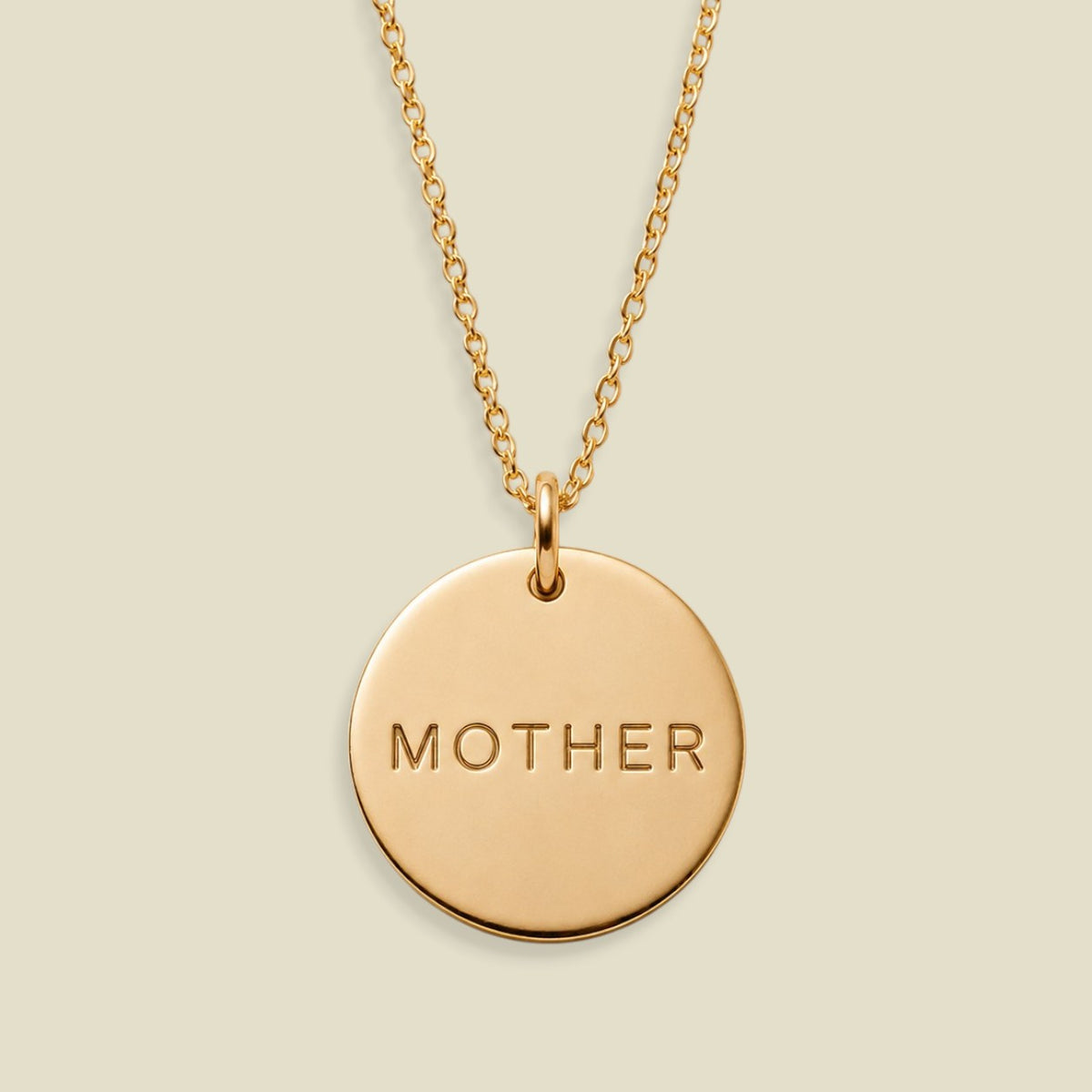 A gold round pendant necklace engraved with the word 'MOTHER' in capital letters, displayed against a solid light beige background.