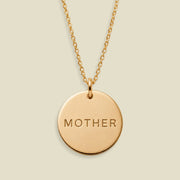 A gold round pendant necklace engraved with the word 'MOTHER' in capital letters, displayed against a solid light beige background.