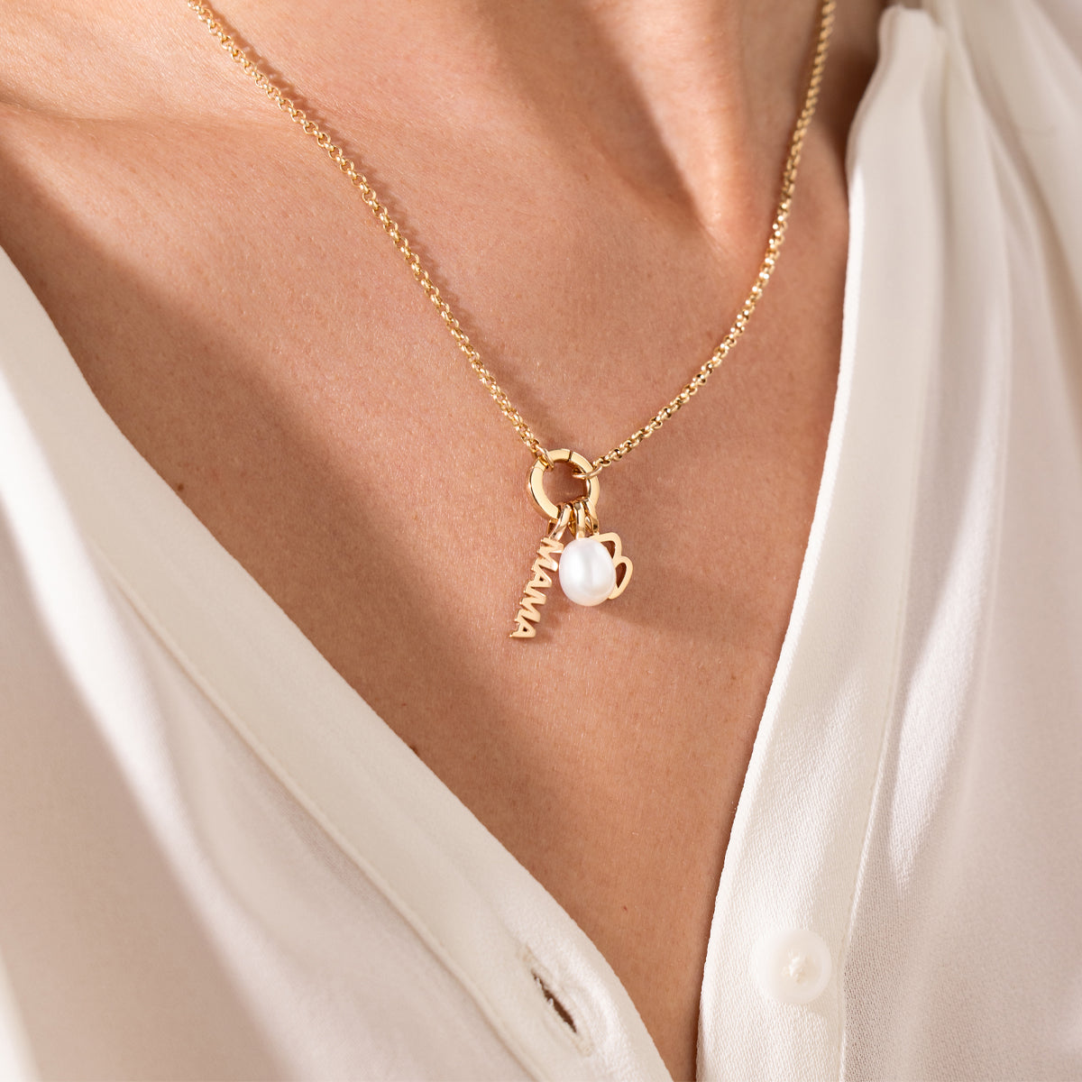 A person wearing a gold necklace with a pearl charm, script font initial 'B' charm and 'MAMA' vertical charm with a white shirt.