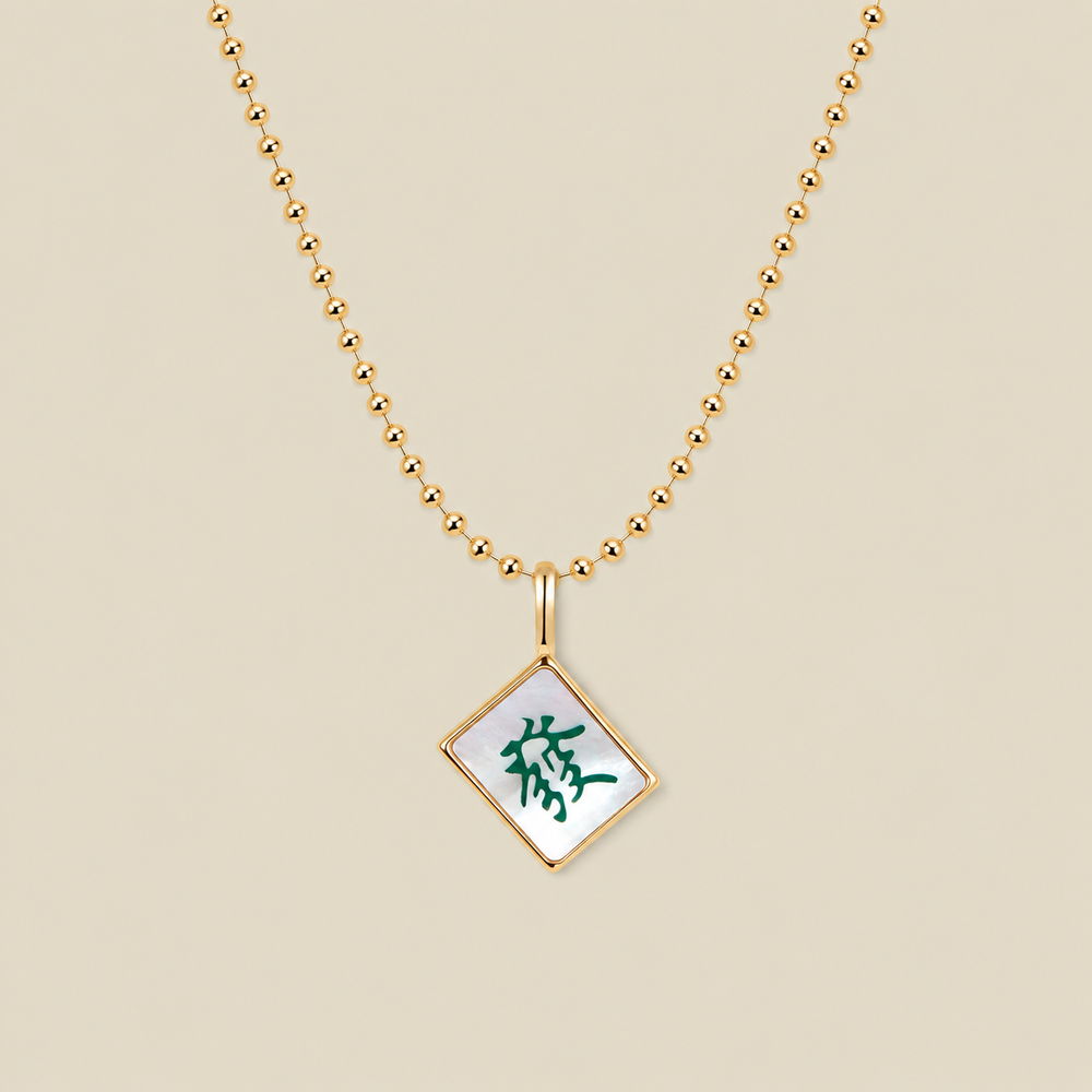 A gold ball chain necklace with a diamond-shaped mahjong tile pendant featuring a white inlay and a green Chinese character, displayed on a solid beige background.