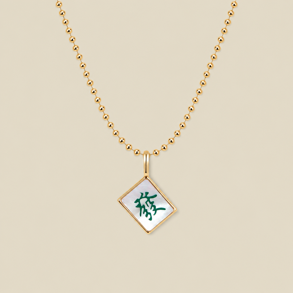 A gold ball chain necklace with a diamond-shaped mahjong tile pendant featuring a white inlay and a green Chinese character, displayed on a solid beige background.