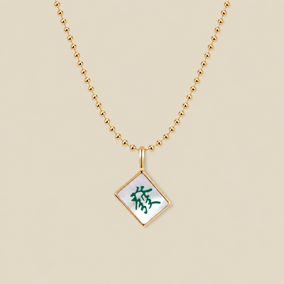 A gold ball chain necklace with a diamond-shaped mahjong tile pendant featuring a white inlay and a green Chinese character, displayed on a solid beige background.