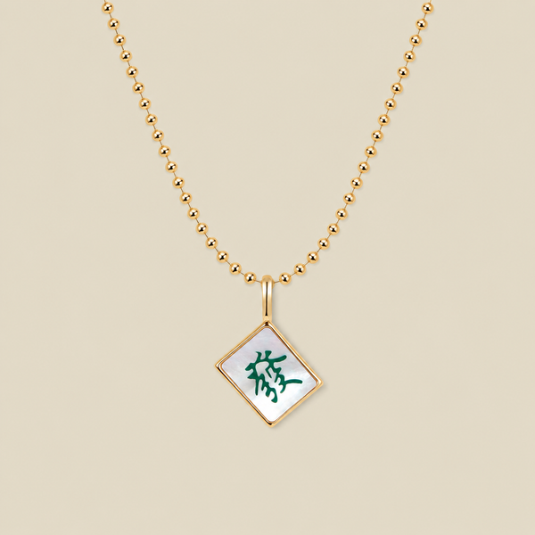 A gold ball chain necklace with a diamond-shaped mahjong tile pendant featuring a white inlay and a green Chinese character, displayed on a solid beige background.