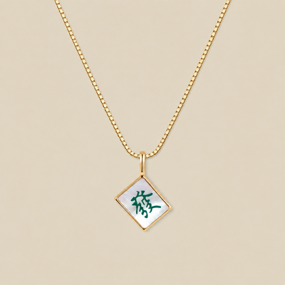 A gold box chain necklace with a diamond-shaped mahjong tile pendant featuring a white mother-of-pearl background and a green Chinese character, displayed on a solid beige background.