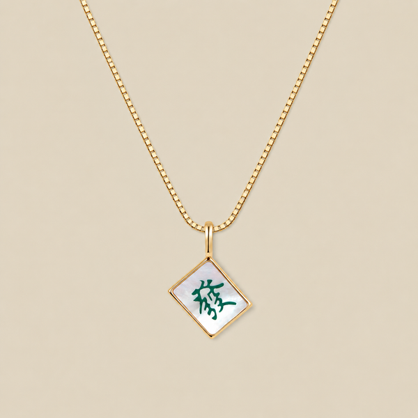 A gold box chain necklace with a diamond-shaped mahjong tile pendant featuring a white mother-of-pearl background and a green Chinese character, displayed on a solid beige background.