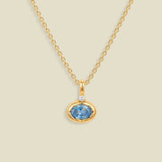 A gold chain necklace with a pendant featuring a small clear stone and an oval bezel set light blue gemstone below it, set against a solid beige background.