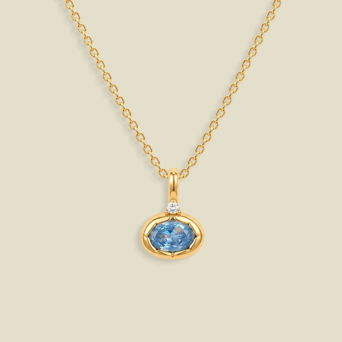 A gold chain necklace with a pendant featuring a small clear stone and an oval bezel set light blue gemstone below it, set against a solid beige background.