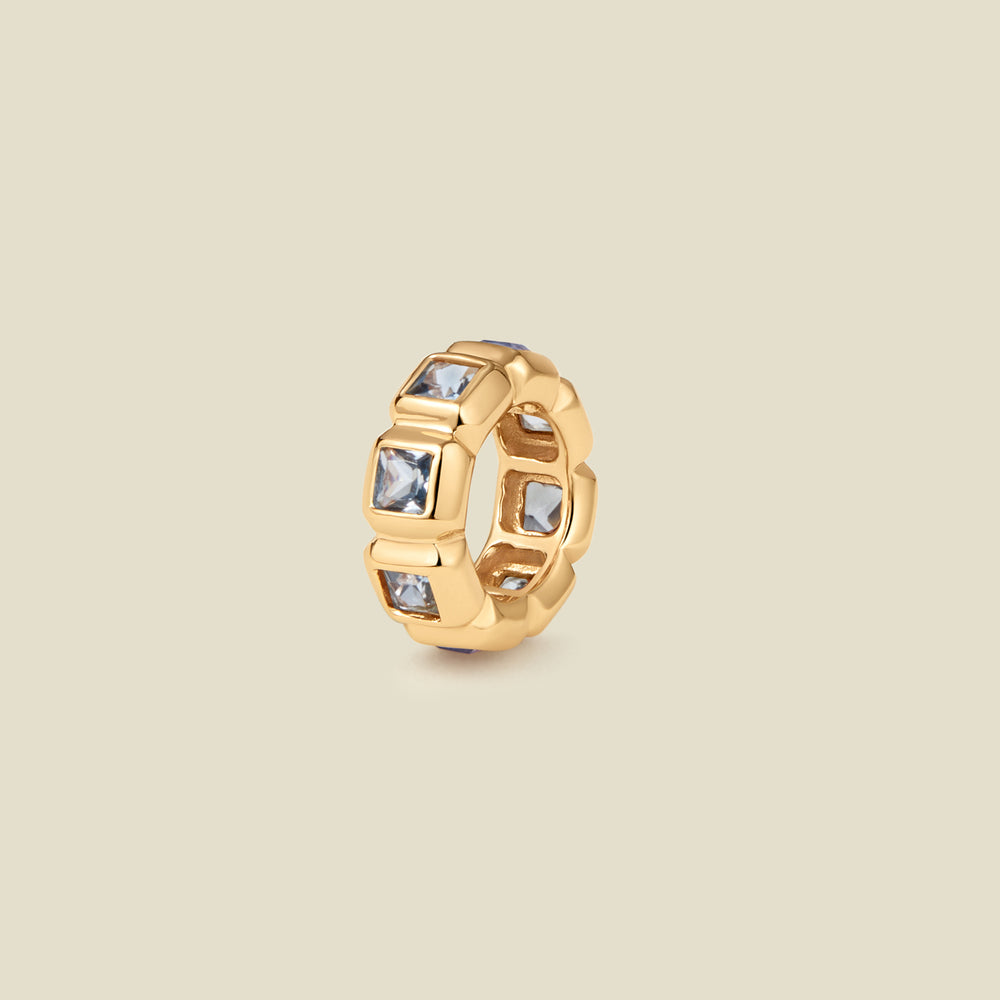 A gold ring charm with multiple bezel set square-cut light grey-blue gemstones set side by side to create the band, shown on a solid beige background.