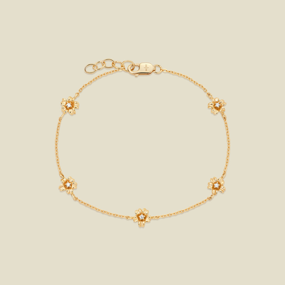 A delicate gold chain bracelet featuring five ornate, textured gold cherry blossom charms with small clear stones, arranged in a circle on a light beige background.