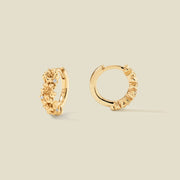 A pair of gold hoop earrings adorned with textured cherry blossom flowers stacked along the hoop, with scattered small clear accent stones, set against a solid beige background.