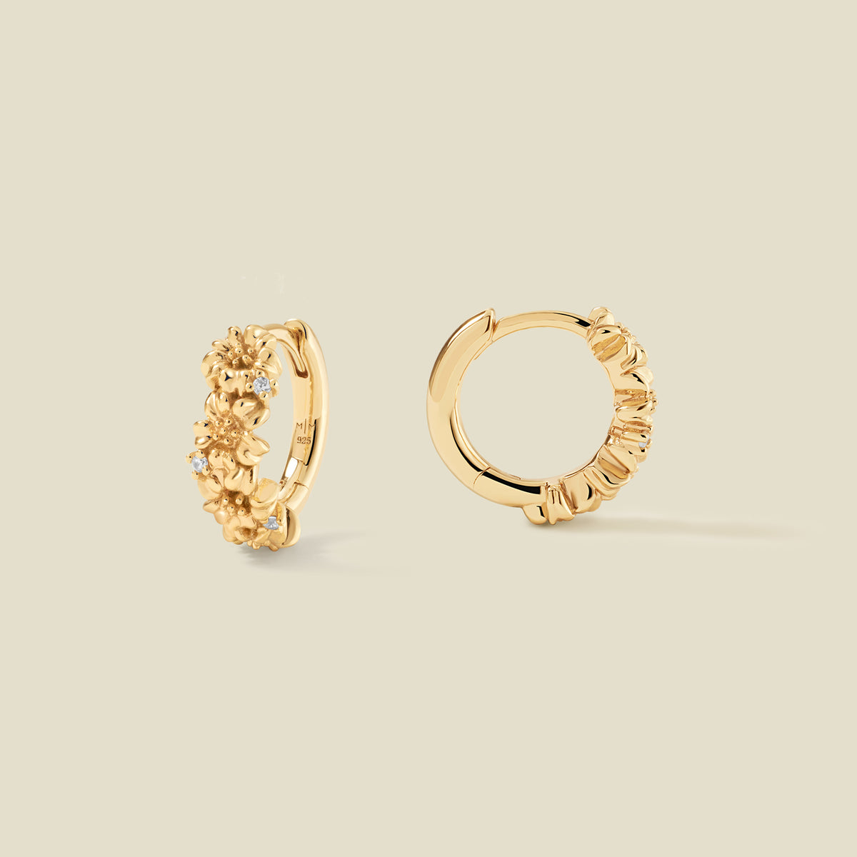A pair of gold hoop earrings adorned with textured cherry blossom flowers stacked along the hoop, with scattered small clear accent stones, set against a solid beige background.