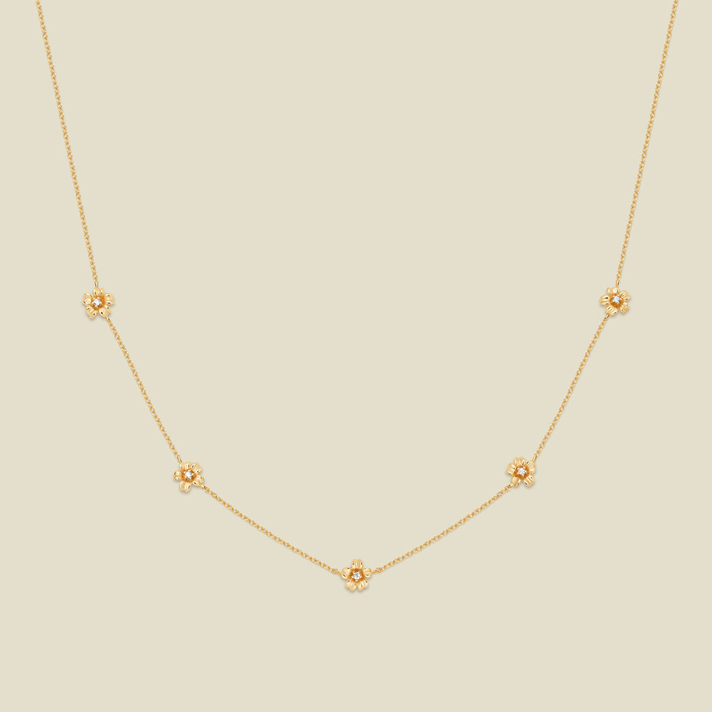 A delicate gold chain necklace featuring five small, evenly spaced textured cherry blossom flower motifs set with tiny clear stones, displayed on a solid light beige background.