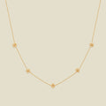 A delicate gold chain necklace featuring five small, evenly spaced textured cherry blossom flower motifs set with tiny clear stones, displayed on a solid light beige background.