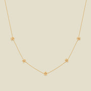 A delicate gold chain necklace featuring five small, evenly spaced textured cherry blossom flower motifs set with tiny clear stones, displayed on a solid light beige background.