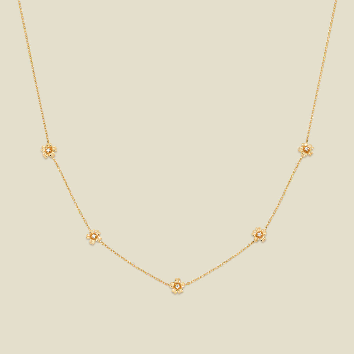 A delicate gold chain necklace featuring five small, evenly spaced textured cherry blossom flower motifs set with tiny clear stones, displayed on a solid light beige background.