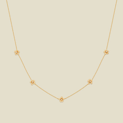 A delicate gold chain necklace featuring five small, evenly spaced textured cherry blossom flower motifs set with tiny clear stones, displayed on a solid light beige background.
