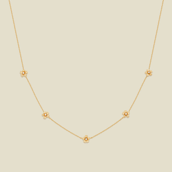 A delicate gold chain necklace featuring five small, evenly spaced textured cherry blossom flower motifs set with tiny clear stones, displayed on a solid light beige background.