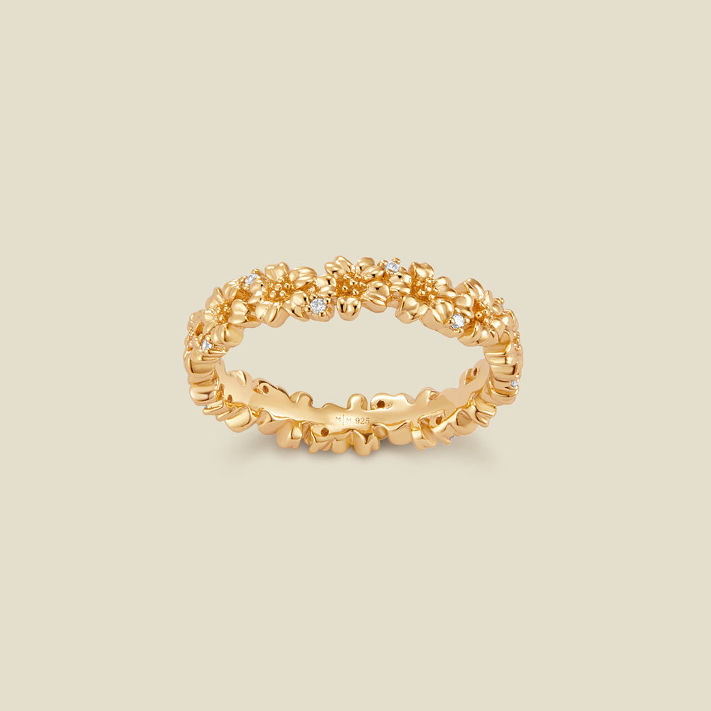 A gold floral-etched ring band with textured cherry blossom flowers and small clear gemstones, shown on a plain warm beige background. 