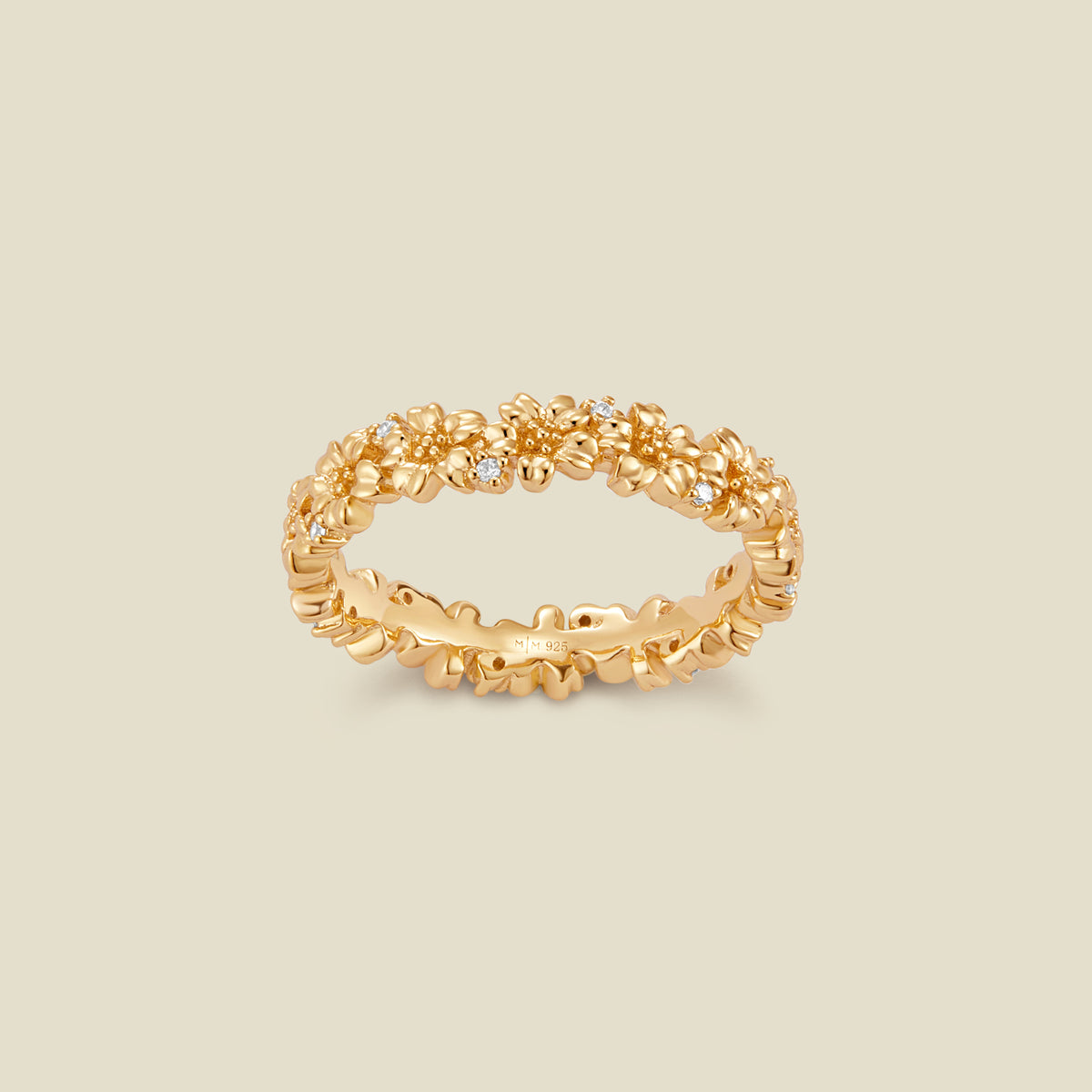 A gold floral-etched ring band with textured cherry blossom flowers and small clear gemstones, shown on a plain warm beige background. 