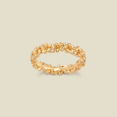 A gold floral-etched ring band with textured cherry blossom flowers and small clear gemstones, shown on a plain warm beige background. 