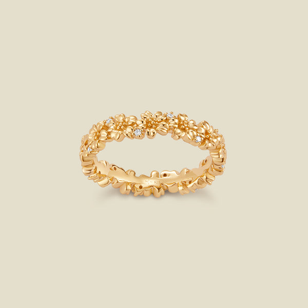 A gold floral-etched ring band with textured cherry blossom flowers and small clear gemstones, shown on a plain warm beige background. 