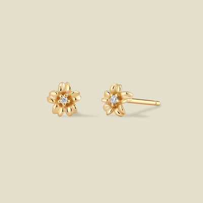 A pair of gold cherry blossom flower stud earrings, one shown from the front with a small clear gemstone in the center and the other from the side showing the post, set against a solid light beige background.
