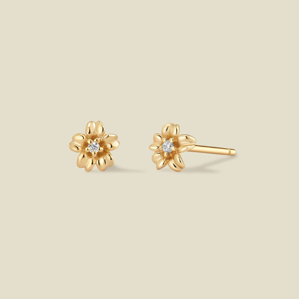 A pair of gold cherry blossom flower stud earrings, one shown from the front with a small clear gemstone in the center and the other from the side showing the post, set against a solid light beige background.