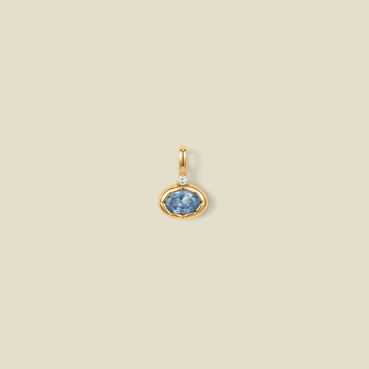 A gold oval pendant charm with a light blue bezel-set oval gemstone and a clear accent stone on top, set against a solid beige background.
