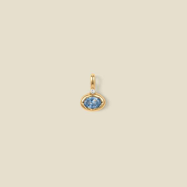 A gold oval pendant charm with a light blue bezel-set oval gemstone and a clear accent stone on top, set against a solid beige background.