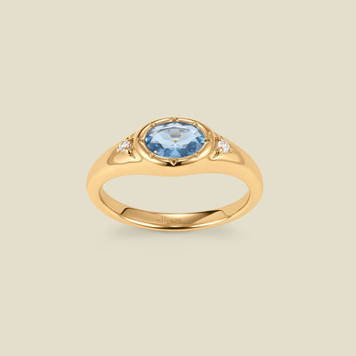 A gold signet ring with a light blue center oval bezel-set gemstone, and two small clear accent stones on either side, set against a solid beige background.
