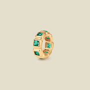 A gold ring charm with multiple bezel set square-cut dark green gemstones set side by side to create the band, shown on a solid beige background.