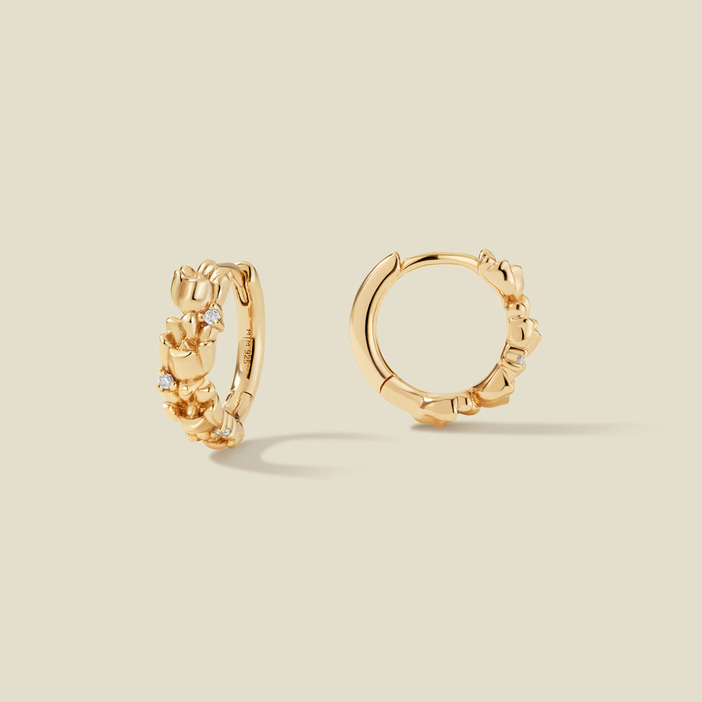 A pair of gold hoop earrings adorned with textured lily of the valley flowers stacked along the hoop, with scattered small clear accent stones, set against a solid beige background.