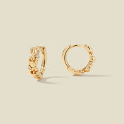 A pair of gold hoop earrings adorned with textured lily of the valley flowers stacked along the hoop, with scattered small clear accent stones, set against a solid beige background.