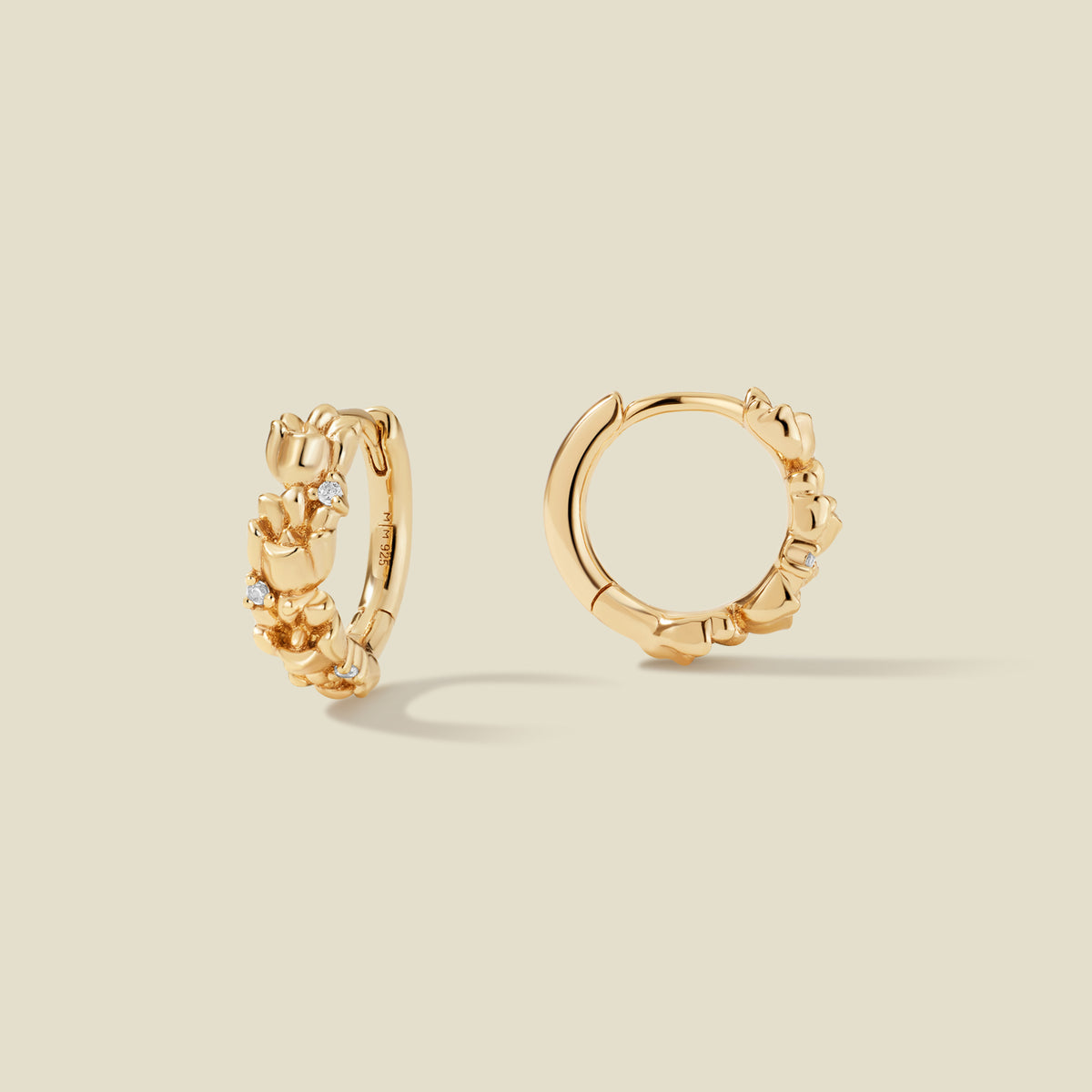 A pair of gold hoop earrings adorned with textured lily of the valley flowers stacked along the hoop, with scattered small clear accent stones, set against a solid beige background.