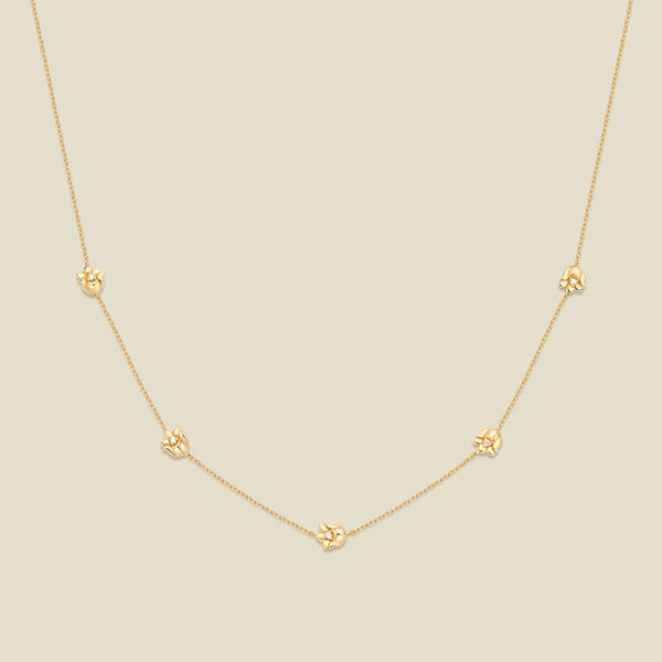 A delicate gold chain necklace featuring five small, evenly spaced textured lily of the valley flower motifs set with tiny clear stones, displayed on a solid light beige background.